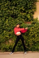 Woman in dynamic dance pose wearing red cropped jacket and black pants against ivy-covered wall