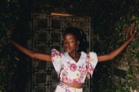 Woman in light blue floral dress with arms outstretched against ornate gate surrounded by ivy