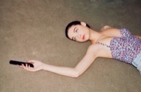 Young woman lying on gray carpet in colorful floral top, holding phone and looking up at camera