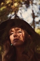 Woman with wavy hair and wide-brimmed hat, dappled sunlight through pine branches on face