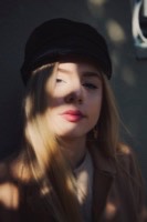 Blonde woman in black newsboy cap and brown jacket with dramatic shadows across face