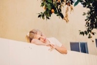 Blonde woman resting chin on hands against white fence with orange tree branches overhead